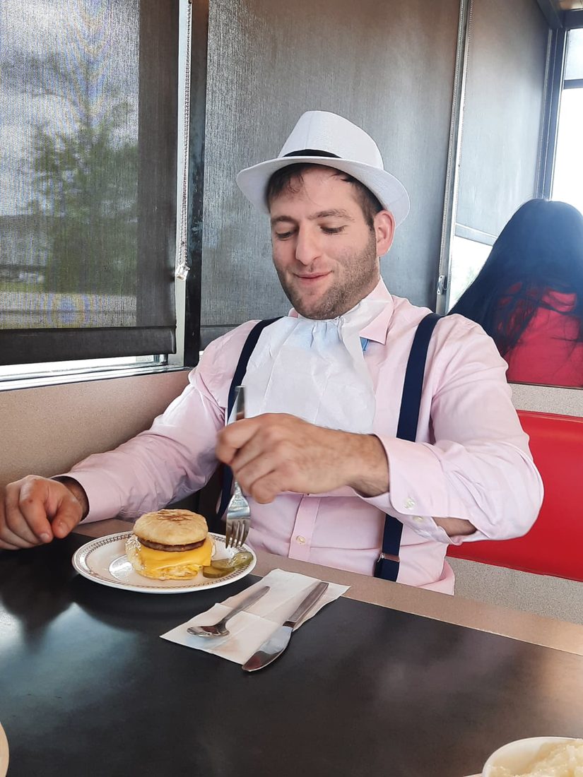 Someone in a hat and bib about to eat a breakfast sandwich at a diner